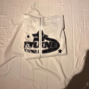 Vit hoodie med svart 4tune-tryck - Säljer en vit hoodie från 4tune med svart tryck framtill och spiraltryck på ryggen. Tröjan har dragkedja, långa ärmar och ribbade muddar. Perfekt för dig som gillar streetwear.