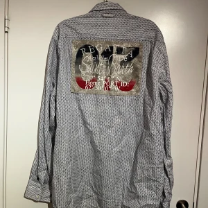 Mönstrad långärmad skjorta från Geographical Norway - Säljer en vit och blå mönstrad skjorta från Geographical Norway med broderade detaljer och tryck på ryggen. Skjortan har klassisk krage, knappar framtill och patchar på ärmen. 