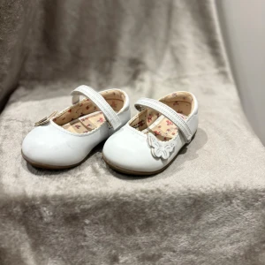 Vita ballerinaskor med fjärilsdetalj Next - Supersöta vita ballerinaskor från Next med kardborreband och en fin fjärilsdetalj på tån. Insidan har blommigt foder och sulan är dekorerad med stjärnor för extra grepp. Perfekta för små fötter som vill sticka ut lite extra.