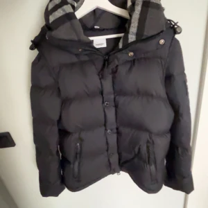 Svart dunjacka Burberry med avtagbara ärmar - Svart pufferjacka från Burberry med avtagbara ärmar, så du kan bära den som väst. Jackan har huva med grå- och svart-rutigt foder, dragkedja och knappar framtill samt två fickor med dragkedja. Stor Burberry-patch på ärmen ger en clean och lyxig vibe.