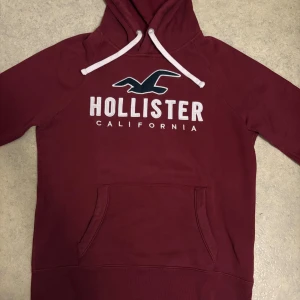 Vinröd hoodie från Hollister M - Snygg vinröd hoodie från Hollister med vit snörning och stor logga på bröstet. Tröjan har huva, magficka och en liten amerikansk flagga på ärmen. Mjuk och skön, perfekt för chill dagar. Passar dig som gillar klassisk streetstyle.