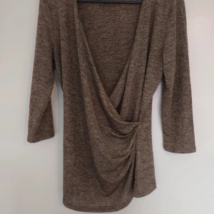 Guldbrun topp med drapering - Snygg guldbrun topp i mjukt stretchigt material med omlott och draperad front. Tröjan har trekvartsärm och djup v-ringning, perfekt för dig som gillar en stilren men ändå cool look.