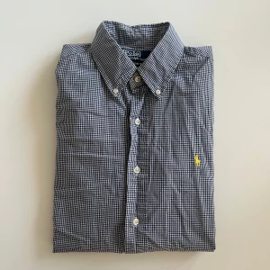 Rutig skjorta från Polo Ralph Lauren - Klassisk rutig skjorta från Polo Ralph Lauren i blått och vitt med gul broderad logga på bröstet. Skjortan har lång ärm, knappar framtill och button down-krage. Tillverkad i mjuk bomull och har en snygg custom fit-passform.                     Mått; bredd: 44cm, längd: 70cm