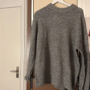 Grå stickad tröja från 157 - Mysig grå stickad tröja från 157 i storlek M. Tröjan har en enkel design med ribbad hals och långa ärmar, perfekt för att hålla dig varm under kyliga dagar. Passformen är loose och materialet känns mjukt och skönt mot huden.