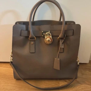 Brun handväska från Michael Kors - Snygg brun handväska från Michael Kors i skinn med guldfärgade detaljer och kedjor på sidorna. Väskan har både handtag och axelrem samt ett ikoniskt MK-lås framtill. Perfekt för dig som vill ha en stilren och lyxig accessoar.