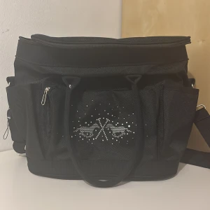 Svart kylväska med strassdetalj - Cool svart kylväska med flera fack, axelrem och handtag. Utsidan har en snygg strassdekor med två hästar och korsade ridspön. Insidan är fodrad med silverfärgat isolerande material som håller dryck och snacks kalla. Perfekt för picknick eller utflykt.