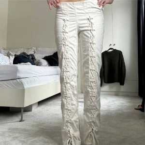 Vita byxor med snörning framtill - Säljer ett par vita byxor med snygg snörningsdetalj längs hela framsidan av benen. Byxorna har raka ben och är tillverkade i ett bomullsliknande material. De har en cool och unik look som sticker ut och passar perfekt till en trendig streetstyle.