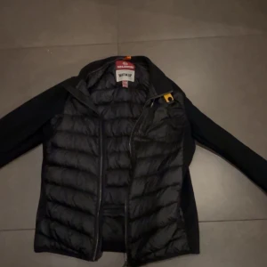 Svart pufferjacka Parajumpers Y-M - Svart pufferjacka från Parajumpers med quiltad front och rygg, samt släta ärmar. Jackan har dragkedja, ikonisk gul detalj vid kragen och patch på ärmen. Materialmix med syntet och polyester, perfekt för kyliga dagar.