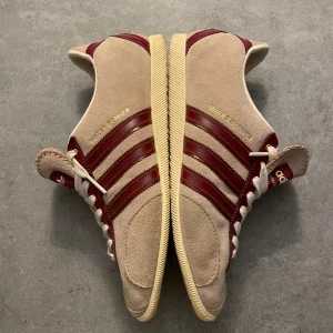  Wales Bonner Japan  beige och vinröd sneakers - Unika Adidas Wales Bonner sneakers i beige mocka med vinröda detaljer och klassiska tre ränder på sidorna. Skorna har vit snörning, rund tå och platt sula. Guldtext på sidan och Adidas-logga på hälen. Snygg retrostil med kontrasterande färger och material.