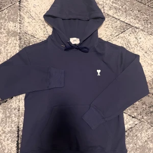 Mörkblå hoodie från Ami Paris - Snygg mörkblå hoodie från Ami Paris med klassisk huva och snörning. Tröjan har ett litet vitt hjärta med A-bokstav broderat på bröstet och en stor magficka. Tillverkad i mjuk bomull, perfekt för chill dagar. Stilren och enkel design. Denna är storlek M men kan även passa folk som bär L.