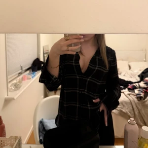 Svart rutig skjorta med v-ringning - Snygg svart skjorta med vitt rutmönster. Skjortan har långa ärmar och en loose passform, perfekt för en chill och stilren look. Materialet känns lätt och flowy, vilket gör den skön att bära hela dagen.