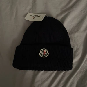 Svart Moncler mössa  - Svart ribbstickad mössa från Moncler med klassisk rundad form och uppvikt kant. Framtill sitter Monclers ikoniska broderade logga i vitt, blått och rött. Perfekt för dig som vill ha en clean och stilren look med exklusiv känsla.