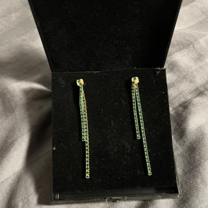 Glittriga gröna och guld örhängen - Säljer ett par långa, glittriga örhängen med två rader gröna stenar infattade i guldfärgad metall. Örhängena har en stilren och modern design som ger en snygg touch till din look.  I guldpläterad stål. Aldrig använda