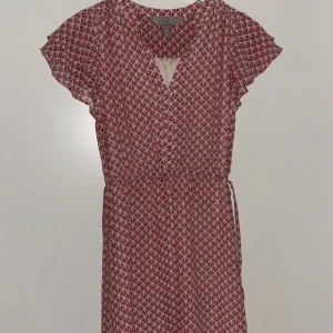 Mönstrad rödvit klänning från H&M - Kortklänning från H&M i rött och vitt med grafiskt mönster. Klänningen har korta ärmar, resår i midjan och knytband i sidan. Lätt och luftig modell med v-ringning och volangdetaljer vid ärmarna. Perfekt för en trendig och avslappnad look.
