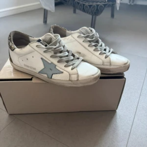 Golden Goose vita sneakers med glitter - Säljer ett par vita Golden Goose sneakers med grå snörning, ljusblå stjärndetalj på sidan och glittrig guldhäl. Skorna har platt sula och rund tå, tillverkade i skinn med slitna detaljer för en cool look. Perfekta för dig som gillar att sticka ut.