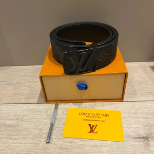 Louis Vuitton svart skinnbälte - Säljer ett stilrent svart Louis Vuitton-bälte i skinn med klassiskt monogrammönster och en stor LV-logga i metall som spänne. Bältet är brett och har en lyxig känsla, perfekt för att lyfta din outfit med en exklusiv touch.