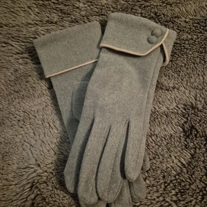 Grå ullhandskar med dekorativa knappar - Snygga grå handskar i mjuk ull med breda muddar och två dekorativa knappar på ovansidan. Diskret beige kant ger en stilren touch. Perfekta för dig som vill ha både värme och stil under kyliga dagar.