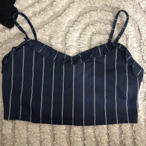 Marinblå randig linnetopp från Lefties - Säljer en marinblå linnetopp från Lefties i storlek M. Toppen har smala axelband, vita vertikala ränder och en liten volangdetalj längs halsringningen. Baksidan har smock för extra komfort och passform. Perfekt för varma dagar och snygg att styla med jeans eller kjol.