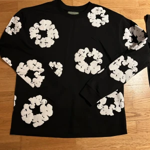 Svart långärmad tröja Denim Tears - Svart långärmad tröja från Denim Tears med stora vita blommiga tryck över hela plagget. Tröjan har rund hals och är tillverkad i mjuk bomull. Perfekt för dig som gillar streetwear och unika prints. ⚠️1:1⚠️
