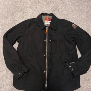 Svart overshirt från Parajumpers - Svart overshirt från Parajumpers med klassisk krage, bröstficka med knapp och patch på ena ärmen. Jackan har en snygg asymmetrisk dragkedja och detaljer i orange. Insidan är grå och har flera etiketter. Perfekt för dig som gillar stilrena och funktionella plagg.