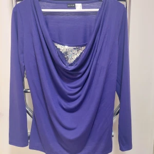 Lila långärmad topp med paljetter - Snygg lila långärmad topp från Body Flirt med draperad front och glittriga paljetter vid halsringningen. Materialet känns mjukt och skönt mot huden. Från rök och djurfritt hem.
