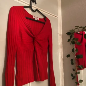 Röd ribbad långärmad topp från New Yorker  - Säljer en snygg röd långärmad topp från New Yorker med ribbad struktur och twistad detalj framtill. Toppen har v-ringad hals och är figurnära i modellen, perfekt för dig som gillar färgstarka plagg med coola detaljer.