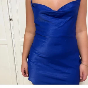 Kornblå satin klänning med slits - Supersnygg kornblå kort klänning i satin med tunna justerbara axelband, draperad urringning och liten slits på sidan. Sitter figurnära och har öppen rygg för en trendig look. Perfekt för dig som vill sticka ut med färg och stil.