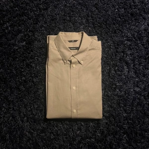 Beige skjorta från J.Lindeberg - Stilren beige skjorta från J.Lindeberg med klassisk krage och knappar framtill. Skjortan är tillverkad i bomull och har en clean look som passar perfekt till jeans eller chinos. Enkel design utan mönster, perfekt för dig som gillar minimalistisk stil.