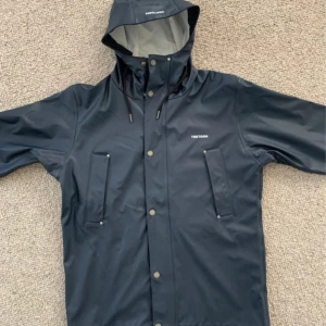 Mörkblå regnjacka från Tretorn XS. Nypris 1000:- - Snygg mörkblå regnjacka från Tretorn i storlek XS. Jackan har huva med snörning, tryckknappar framtill och två stora fickor. Perfekt för regniga dagar och har en stilren look med diskret logga på bröstet.