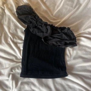 Svart oneshoulder topp med volang - Säljer en svart oneshoulder topp med stor volangdetalj över axeln. Toppen är tight och har en cool asymmetrisk design som ger en trendig look. Perfekt för dig som vill sticka ut med en unik och stilren topp.