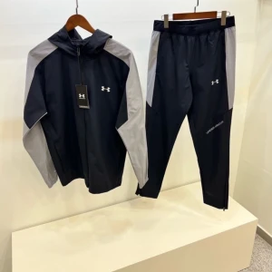 Under Armour svart träningsset - Snyggt träningsset från Under Armour med svart jacka och byxor, båda med grå paneler på sidorna. Jackan har huva och dragkedja framtill, och byxorna har logga på benet. Perfekt för dig som gillar sportig stil och vill ha något bekvämt till gymmet.