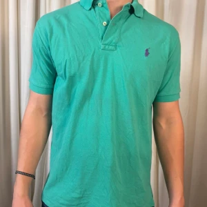 Grön pikétröja från Polo Ralph Lauren - Snygg grön pikétröja från Polo Ralph Lauren i klassisk modell med krage och två knappar. Tröjan har korta ärmar och den ikoniska lilla logon broderad på bröstet i lila. Tillverkad i mjuk bomull som är skön mot huden. Perfekt för dig som gillar stilren och sportig look.