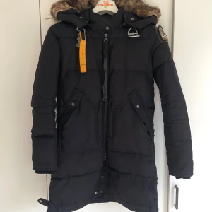 Svart Parajumpers dunjacka med päls - Svart lång dunjacka från Parajumpers med stor huva och avtagbar päls. Jackan har ikonisk gul rem med metallspänne, patch på ärmen och två stora fickor framtill. Använd en vinter, i fint skick! OBS! Finns ett litet hål på mindre än 1 mm vid slutet av armen. Syns lite på första bilden men påverkar inte jackans funktion. Skickar fler bilder vid intresse!