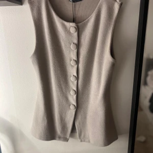 Beige väst med klädda knappar från Zara - Stilren beige väst från Zara med rund halsringning och fem klädda knappar framtill. Västen har en figurnära passform och är tillverkad i ett mjukt material som ger en clean look. Perfekt att styla över en skjorta eller t-shirt för en trendig vibe.