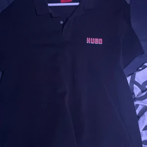 Svart pikétröja från Hugo - Svart pikétröja från Hugo med röd logotyp broderad på bröstet. Klassisk krage och knappslå framtill. Tröjan har en clean och stilren look, perfekt för dig som gillar enkel design med en touch av branding. Äkta Hugo boss köpt för 1900