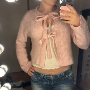 Ljusrosa stickad kofta med rosetter - Supergullig ljusrosa stickad kofta från Gina tricot med två stora rosetter framtill som knytning. Koftan är croppad, har långärmade ärmar och ett ribbat mönster. Perfekt för dig som gillar söta detaljer och mjuka material. Man kan knyta upp rosetterna!