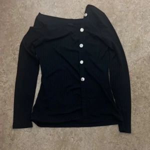Svart offshoulder topp med knappar - Snygg svart offshoulder topp med långa ärmar och  knappar längs framsidan. Toppen har ett ribbat mönster och sitter tight mot kroppen. Perfekt för dig som gillar en stilren look med lite edge.