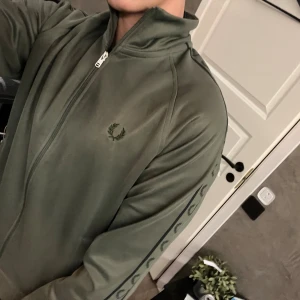 Olivgrön track jacket Fred Perry S - Snygg olivgrön track jacket från Fred Perry med dragkedja och ribbade muddar. Tröjan har Fred Perry-logga på bröstet och längs ärmarna, samt ståkrage. Materialet är mjukt och syntetiskt, perfekt för en sportig och chill stil. Skick 7/10