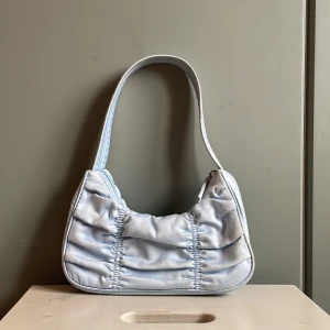 INTRESSEKOLL babyblå axelväska  - !!!INTRESSEKOLL!!!Supersöt ljusblå axelväska från Monki i glansigt satinmaterial. Väskan har rynkade detaljer på framsidan och en praktisk dragkedja upptill. Insidan är fodrad och har ett extra fack för småsaker. Väskan ser mer babyblå ut än på bilderna. Den är lite nopprig🩵🩵