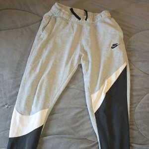 Grå mjukisbyxor från Nike med snörning - Snygga grå mjukisbyxor från Nike med svart och vit panel på benen. Byxorna har elastisk midja med svart snörning, sidofickor och Nike-logga på vänster lår. Perfekta för chill eller träning, tillverkade i mjukt bomullsmaterial.