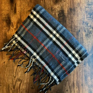 Burberry Halsduk - Klassisk Burberrys-halsduk i 100% lammull, tillverkad i England. Mjukt och varmt material i ikoniskt rutmönster. Passar både dam och herr. I mycket bra skick utan fläckar eller hål – perfekt för höst och vinter.