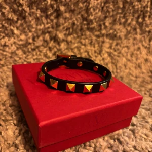 Valentino Garavani armband  - Säljer ett svart armband med guldiga pyramidnitar från Valentino Garavani. Armbandet har justerbar spänne. Allt kommer med original ask, påse och certifikat. Extra bitar finns med. Nypris runt 3000.