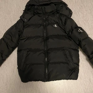 Svart pufferjacka från Calvin Klein - Svart pufferjacka från Calvin Klein med huva och dragkedja framtill. Jackan har CK-logga på bröstet och ärmen, samt två sidofickor. Puffad design med horisontella sömmar och hög krage. Perfekt för kalla dagar och riktigt snygg streetstil. En liten defekt som visas på sista bilden, skriv vid frågor!