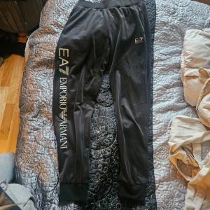 Svarta mjukisbyxor EA7 Emporio Armani - Svarta mjukisbyxor från EA7 Emporio Armani med bred resår i midjan och muddar vid bensluten. Snyggt tryck med logga längs ena benet i silver. Tillverkade i mjukt material som känns skönt mot huden. Perfekta för chill eller träning. Enda skavanken är att dom är lite noppriga.