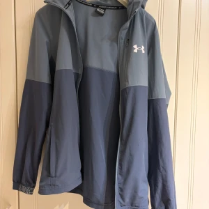 Blå vindjacka från Under Armour - Snygg tvåtonad blå vindjacka från Under Armour med huva och vit logga på bröstet. Jackan har dragkedja framtill, elastiska muddar med tryckt text och en sportig look. Perfekt för dig som gillar streetstyle och funktionella plagg. Priset kan diskuteras vid snabb affär
