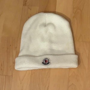 Vit Moncler mössa i ull - Snygg vit mössa från Moncler med klassisk uppvikt kant och broderad logotyp framtill. Tillverkad i mjuk ull som håller dig varm under kalla dagar. Enkel och stilren design som passar perfekt till vinterlooken.