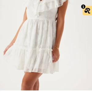 Vit kort volangklänning med v-ringning - Superfin vit kort klänning med volanger och v-ringning. Klänningen har ett subtilt blommönster i tyget, korta ärmar och har en flowy kjol med volangkant. Perfekt för student. Använd en gång men syns inte att den är använd. Öppen för prisförslag