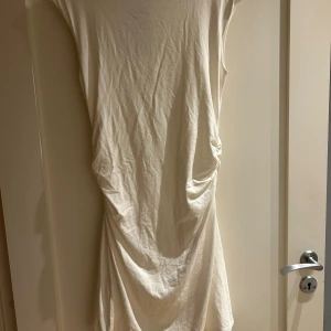 Vit klänning från Bik Bok - En vit klänning från Bik Bok i mjuk bomull. Klänningen har ärmlös design och snygg drapering på sidan som ger en cool omlott-effekt. Perfekt för sommarens alla tillfällen. Enkel och stilren modell som är lätt att matcha.