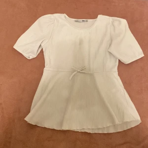 Vit blus med rosett från 157 - Säljer en söt vit blus från 157 med korta puffärmar och rund hals. Blusen har en liten rosett i midjan och är tillverkad i ett ribbat bomullstyg som ger en mjuk känsla. Passformen är normal och nederdelen är lätt utsvängd för en feminin look.