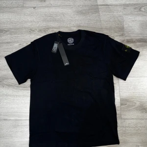 Svart t-shirt från Stone Island - Svart t-shirt från Stone Island med klassisk rund hals och korta ärmar. Ikonisk Stone Island patch sitter på vänster ärm. T-shirten är gjord i mjuk bomull och har en clean, stilren look som passar perfekt till streetwear. Taggarna sitter kvar. Ete